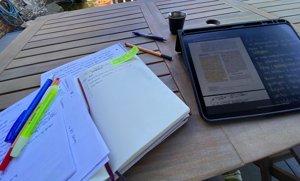Notizbuch, Zettel und Stifte neben Kaffeebecher und iPad auf einem Gartentisch
