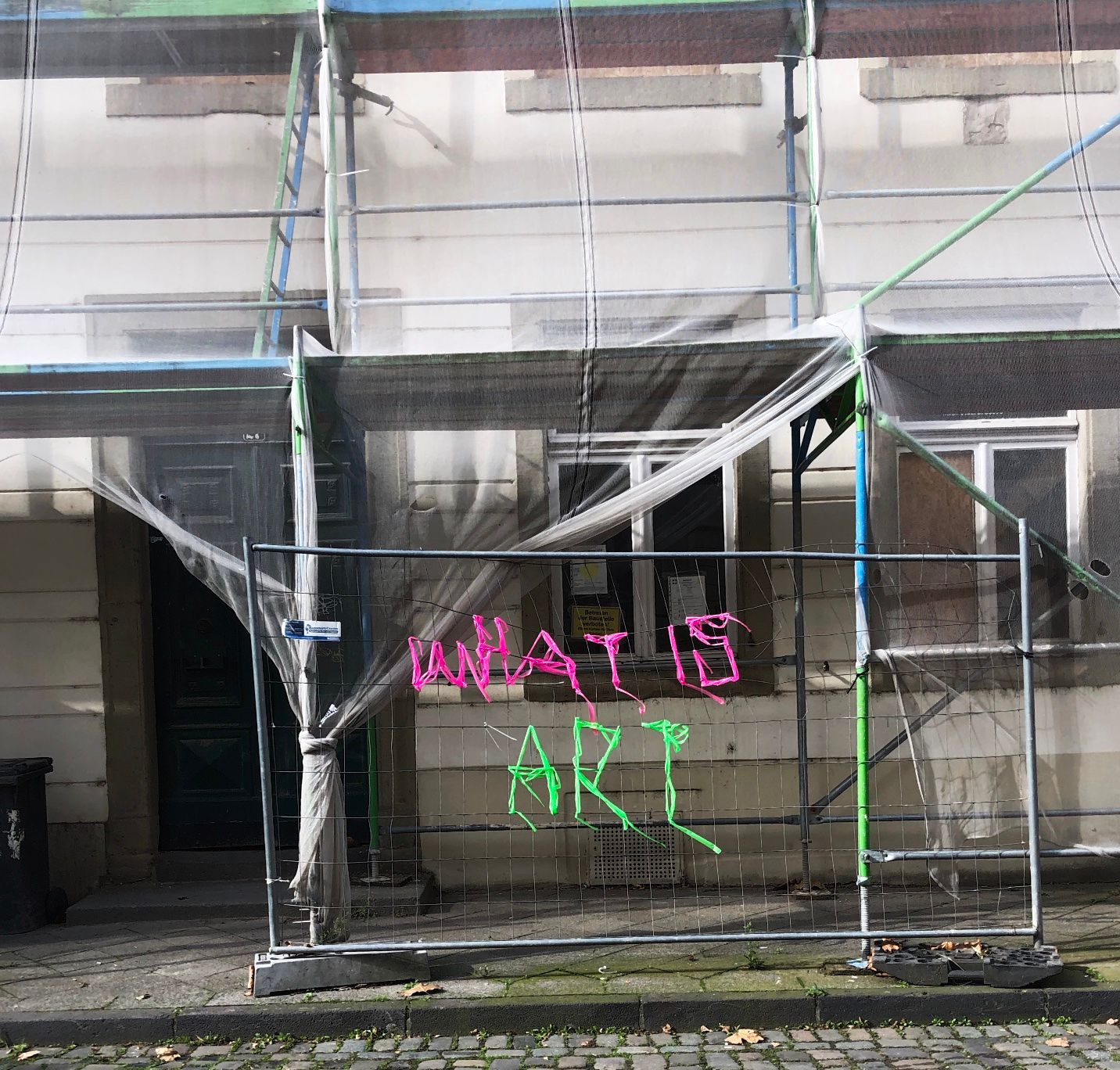 Bauzaun vor einer Hausfassade. Auf dem Zaun wurde mit buntem Band "What is Art" geschrieben.